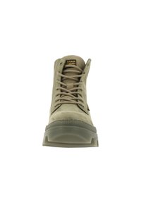 Vue de face d’une botte montante beige à lacets avec une semelle en caoutchouc épaisse et une étiquette de marque visible sur la languette.