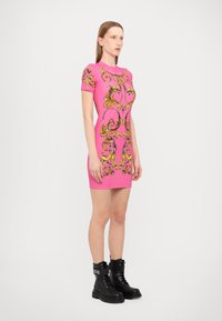 Abito aderente rosa con decorazioni ornate gialle, maniche corte e collo rotondo, abbinato a stivali da combattimento neri alla caviglia.