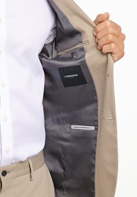 Beige colbert met een grijze satijnen binnenvoering, met een label, een knoop en een horizontale zak aan de linkerkant.