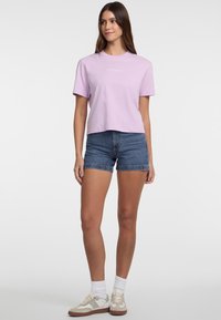 Lillā puķu formas T-krekls ar īsām piedurknēm, ar "GUESS JEANS" logotipu, kombinēts ar zilām džinsa šortiem un bežām kedām ar baltām akcentiem.