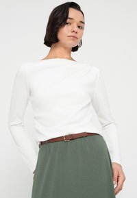 Λευκό τοπ με μακριά μανίκια και φαρδιά λαιμόκοψη, συνδυασμένο με πράσινη φούστα που διαθέτει καφέ ζώνη, κατασκευασμένη από μαλακό, λείο ύφασμα.