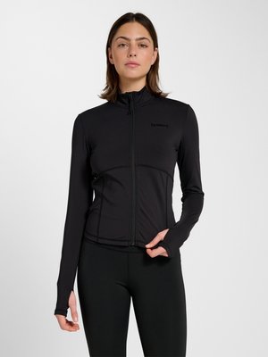 Vrouw draagt een nauwsluitend zwart zip-up top met lange mouwen en duimgaten, staand tegen een egale lichte achtergrond.