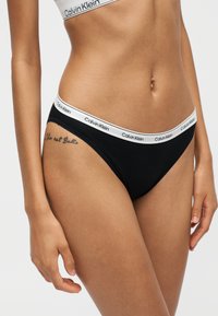 Γυναίκα που φοράει μαύρα εσώρουχα Calvin Klein σε στιλ μπικίνι με λευκή ελαστική μέση με σήμα, ορατό τατουάζ στο ισχίο και ασορτί λευκό αθλητικό σουτιέν.