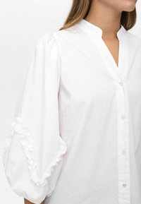 Witte blouse met pofmouwen, gerimpeld randje op de mouwen, Mandarin-kraag en een sluiting met knopen aan de voorkant, gedragen door een persoon die van de nek naar beneden is afgebeeld.