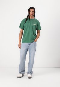Groene katoenen t-shirt met een logo op de borst, gecombineerd met lichtblauwe denim jeans en grijze schoenen. Eenvoudig ontwerp, relaxte pasvorm.