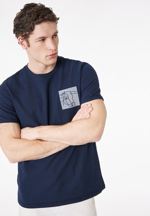 Junger Mann mit lockigem Haar, der ein dunkelblaues T-Shirt mit minimalistischem Kuhgesicht-Design auf dem Brustpatch trägt, Arme verschränkt, schaut nach links.