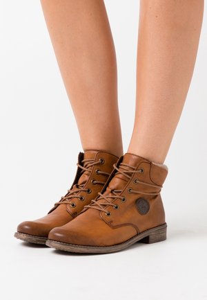 Botas de tobillo de piel marrón con puntera redonda, diseño con cordones y forro interior suave. Presenta un parche con el logo en el lateral y una suela texturizada.