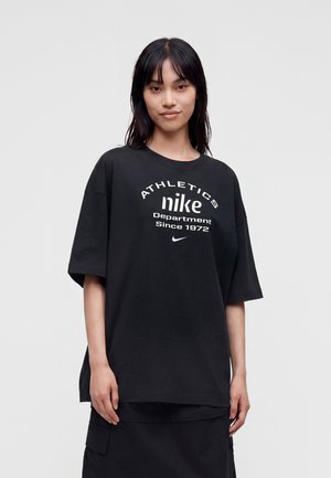 Junge Frau mit glatten dunklen Haaren trägt ein übergroßes schwarzes Nike-T-Shirt mit weißem Athletics-Schriftzug und steht vor einem schlichten hellen Hintergrund.