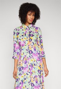 Vestido floral de botões com mangas três quartos, apresentando um fundo verde pastel e padrões vibrantes de flores roxas, amarelas e azuis.