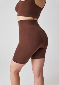 Woman wearing high-waisted brown compression shorts and matching sports bra, shown from side against a plain light background.