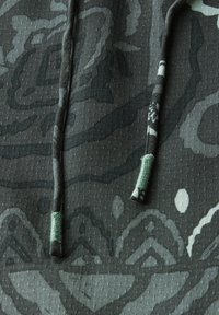 Un tissu à motif camouflage dans des nuances de vert foncé présente une surface texturée, avec des accents plus clairs contrastants et un cordon de serrage avec des embouts verts.