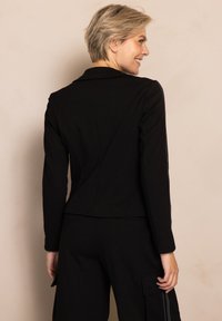 Schwarzer, taillierter Blazer mit langen Ärmeln, Revers mit Kerbkragen und figurbetontem Rücken. Aus glattem Stoff, kombiniert mit schwarzen Cargo-Hosen.