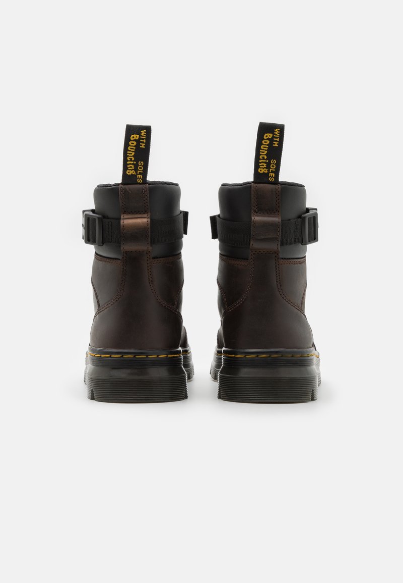 Bruine leren enkelboots met zwarte accenten, een gespband, gele hieltreklussen en een dikke rubberen zool met gele stiksels.