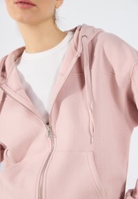 Hoodie rose à fermeture éclair en tissu doux, doté d'une poche kangourou, de quincaillerie en argent pour la fermeture éclair et d'une capuche avec cordon de serrage.