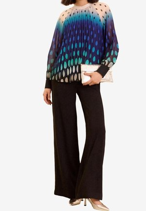 Kvinde iført en plisseret bluse med lange ærmer med blågrønne polkaprikker, sorte vidde bukser, guld clutch og guld spidse hæle.