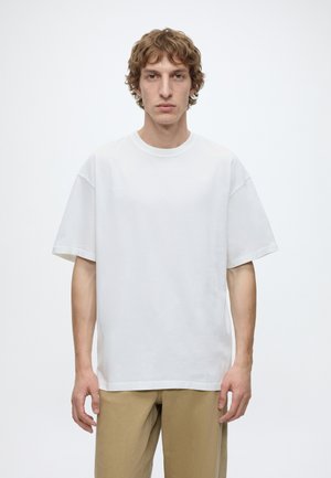 Junger Mann mit welligem hellbraunem Haar trägt ein schlichtes weißes übergroßes T-Shirt und beige Hose, steht vor einem schlichten weißen Hintergrund.