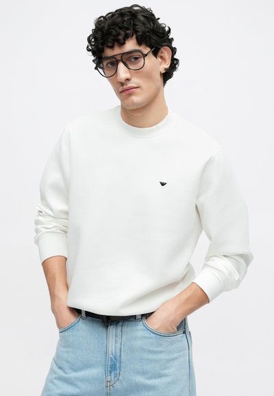 Λευκό φούτερ με στρογγυλή λαιμόκοψη και μακριά μανίκια, με μικρό σκούρο λογότυπο στο στήθος, συνδυασμένο με ανοιχτό μπλε τζιν παντελόνι.