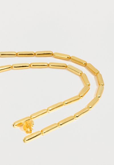 Κολιέ χρυσαφί χρώματος με επιμηκυσμένες κυλινδρικές χάντρες, τοποθετημένες σε χαλαρό βρόχο πάνω σε απλό λευκό φόντο.