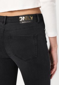 Jeans de denim negro con un diseño de cinco bolsillos. Presenta una etiqueta dorada con el nombre de la marca en la cinturilla. Textura suave y forma ajustada.