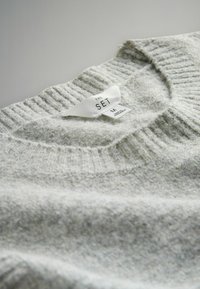 Grau gestrickter Pullover mit ribbelndem Ausschnitt, mit einem weißen Pflegeetikett, das "THE SET" und die Größe "M" angibt. Weicher, strukturierter Stoff.