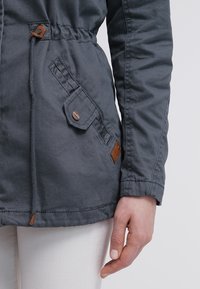 Chaqueta parka gris con cintura fruncida, que presenta un bolsillo con solapa y cierre de botón, detalle en cuero y tejido texturizado.