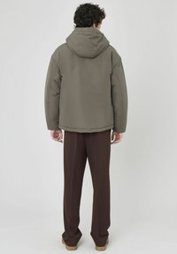 Veste matelassée à capuche de couleur olive unie, avec une coupe décontractée et des poignets élastiques. Portée avec un pantalon marron ample.