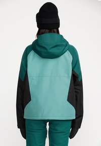 Veste de ski teal et noire avec une capuche, présentant un design coloré en blocs et un tissu texturé. Le modèle porte un bonnet noir.
