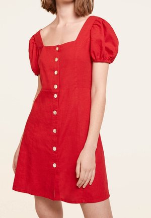 Mujer con un vestido rojo de manga corta con mangas abullonadas y una fila de botones blancos al frente, de pie delante de un fondo liso.