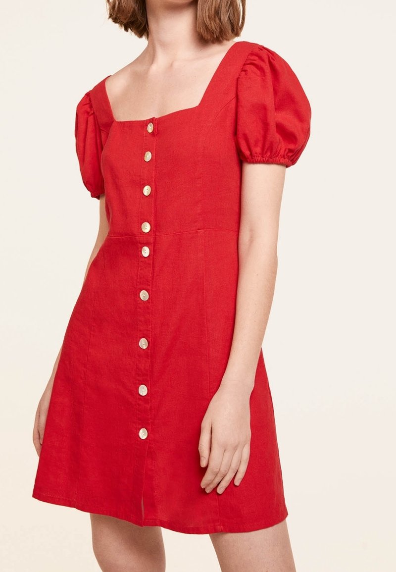 Femme portant une robe rouge à manches courtes bouffantes avec une rangée de boutons blancs à l'avant, debout devant un fond uni.