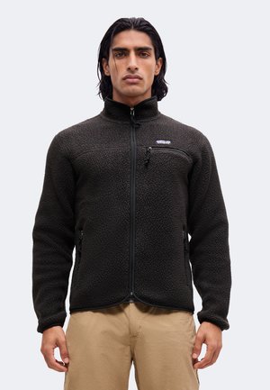 Homme aux cheveux bruns mi-longs portant une veste polaire noire avec des poches zippées et un pantalon beige, debout devant un fond uni.