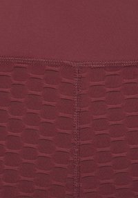 Burgunderfarbene Trainingsleggings mit einem glatten oberen Abschnitt und einem strukturierten unteren Abschnitt, der ein geometrisches Muster aufweist. Sichtbare verstärkte Nähte.