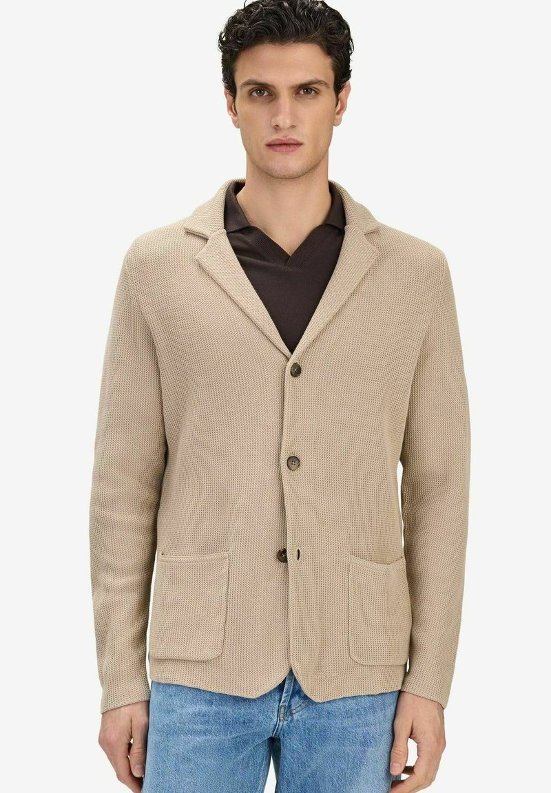 Homme portant un blazer beige en tricot texturé sur une chemise marron foncé et un jean bleu clair, debout devant un fond blanc uni.