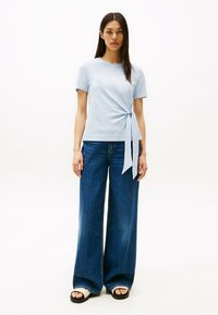 Top azzurro chiaro a maniche corte con dettaglio a nodo laterale, abbinato a jeans in denim blu scuro a gamba larga e sandali neri con cinturini bianchi.