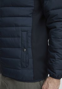 Marineblå puffjakke med horisontal kviltedesign, sidelomm med trykknapp og teksturerte stretchstoffpaneler på sidene.