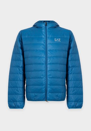 Jeune homme debout portant une doudoune bleue à capuche, un t-shirt noir, un pantalon beige et des baskets blanches sur fond uni.