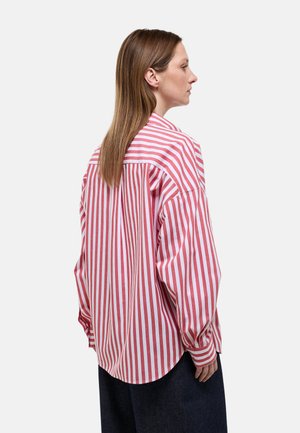 Femme aux cheveux bruns raides portant une chemise ample à rayures verticales rouges et blanches, vue de profil et de dos sur un fond uni.