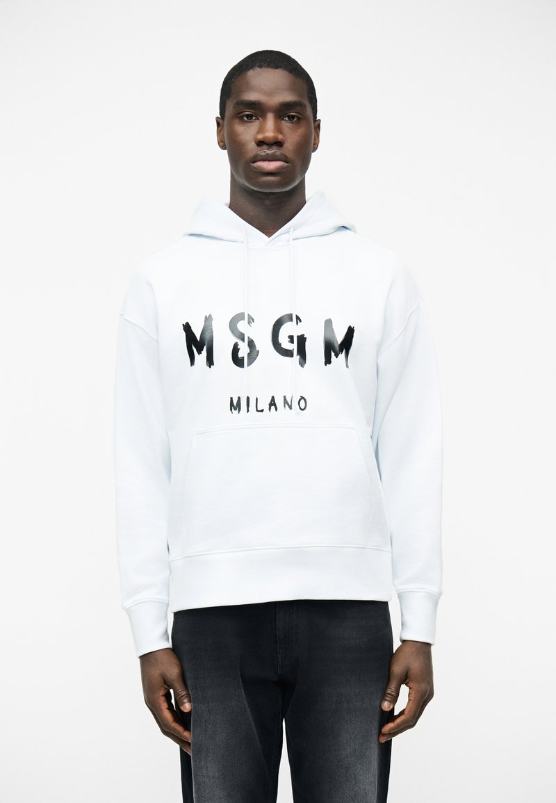 Νέος άνδρας που φοράει λευκό φούτερ MSGM Milano και μαύρα τζιν, στέκεται μπροστά σε απλό λευκό φόντο.
