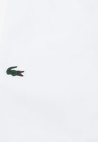 Bijela polo majica od teksturiranog tkanja s izvezenim zeleno-crvenim logotipom na lijevoj strani prsa. Jednostavan, klasičan dizajn.