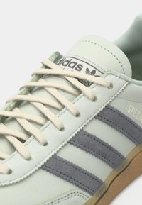 Απαλά πράσινα σουέντ αθλητικά παπούτσια με λευκά κορδόνια, που διαθέτουν τρεις γκρι ρίγες και ετικέτα με λογότυπο στη γλώσσα. Λαστιχένια σόλα με υφή σε λεπτομέρειες.