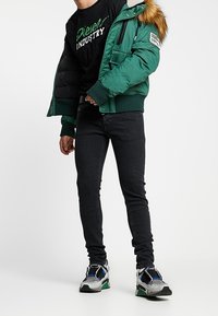 Zelená bombera s kožešinovým lemováním, černé tričko s grafikou, černé úzké džíny a barevné tenisky. Texturovaná tkanina a vrstevnatý design.