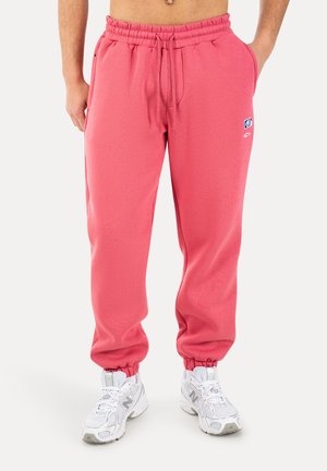 Uomo che indossa pantaloni sportivi color corallo con vita e polsini elasticizzati, abbinati a scarpe bianche New Balance, in piedi contro uno sfondo bianco.