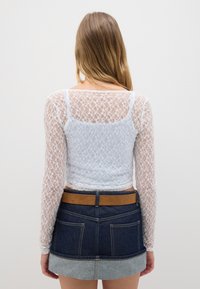 Top lungo a maniche con pizzo bianco, dal design aderente e motivo floreale. Abbinato a una gonna di jeans con una cintura marrone e dettagli sul bordo.