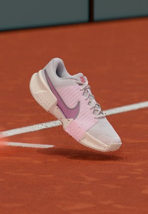 Jasnoróżowy i biały tenisowy but Nike wzbijający kurz z glinianego kortu tenisowego w pobliżu linii końcowej.
