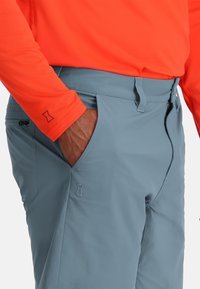 Lichte grijze buitenbroek met een soepele textuur, voorzien van een zijzak en gestikte ontwerpdetails. Gecombineerd met een feloranje shirt.