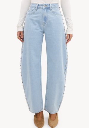 Jean bleu clair à jambes larges avec des clous argentés le long des coutures extérieures, porté avec des chaussures beiges à bouts pointus et un haut blanc à manches longues.