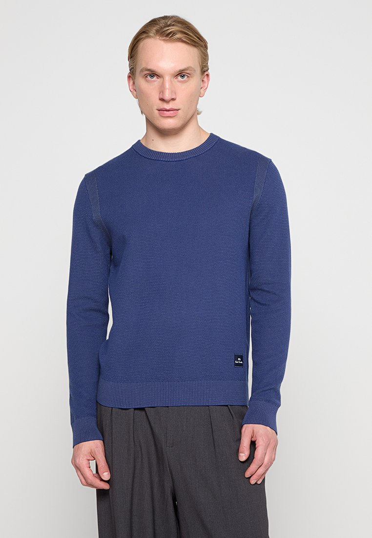 Mörkblå stickad tröja med rund halsringning, ribbade ärmslut och nederkant, med en liten logotag vid midjan. Slät textur, avslappnad passform.