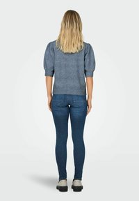 Femme blonde se tenant de dos, portant un pull gris à manches courtes, un jean skinny bleu foncé et des chaussures à plateforme beiges.