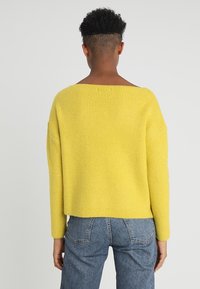 Osoba s krátkými kudrnatými vlasy, oblečená v jasně žlutém žebrovaném svetru a modrých džínech, zobrazená zezadu proti jednoduchému pozadí.