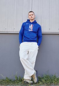 Niebieska bluza Nike z logo NBA, białe spodnie o luźnym kroju i szare buty hikingowe z pomarańczowymi sznurowadłami. Gładkie tekstury i casualowy design.