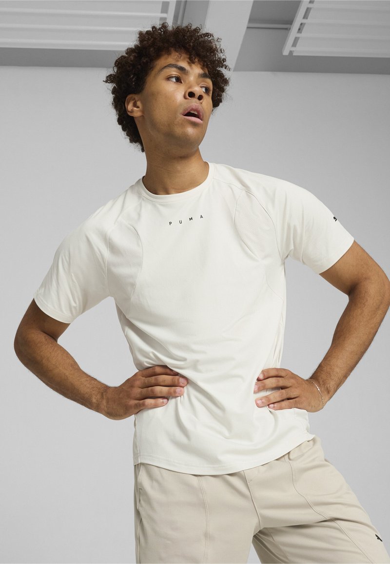 Camiseta deportiva blanca con una textura suave, mangas cortas y un sutil logotipo en el pecho. Combinada con pantalones cortos beige claros.